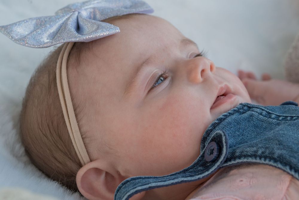 Baby wearing denim with silver bow headband resting on white surface.