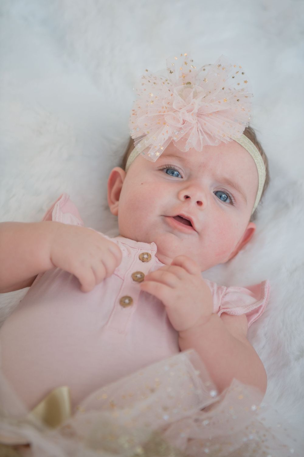 Baby in pink outfit with tulle bow headband lying on white background.