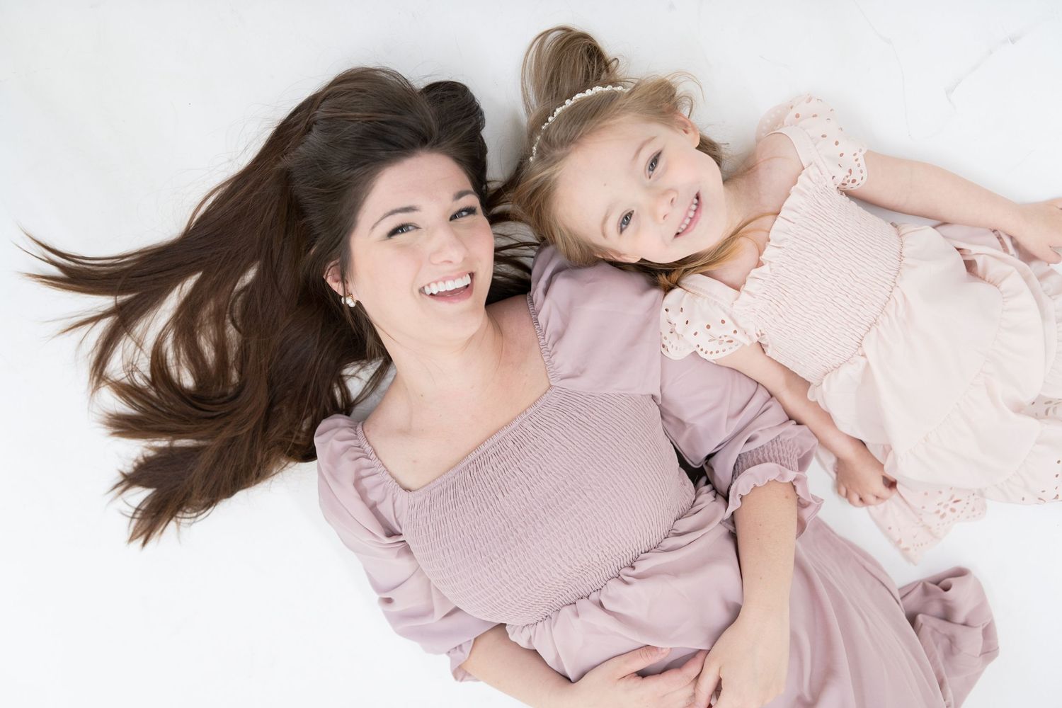 Mother & Daughter laying on white backdrop wearing coordinating pink dresses during photoshoot.