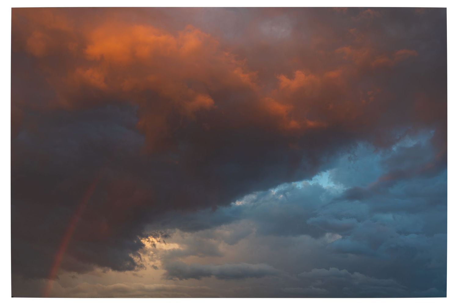 Dramatic stormy sky with vivid orange-red clouds and a rainbow breaking through dark blue storm clouds at sunset.