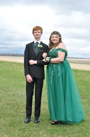 Formal couple poses in black suit and emerald green gown on grassy field.