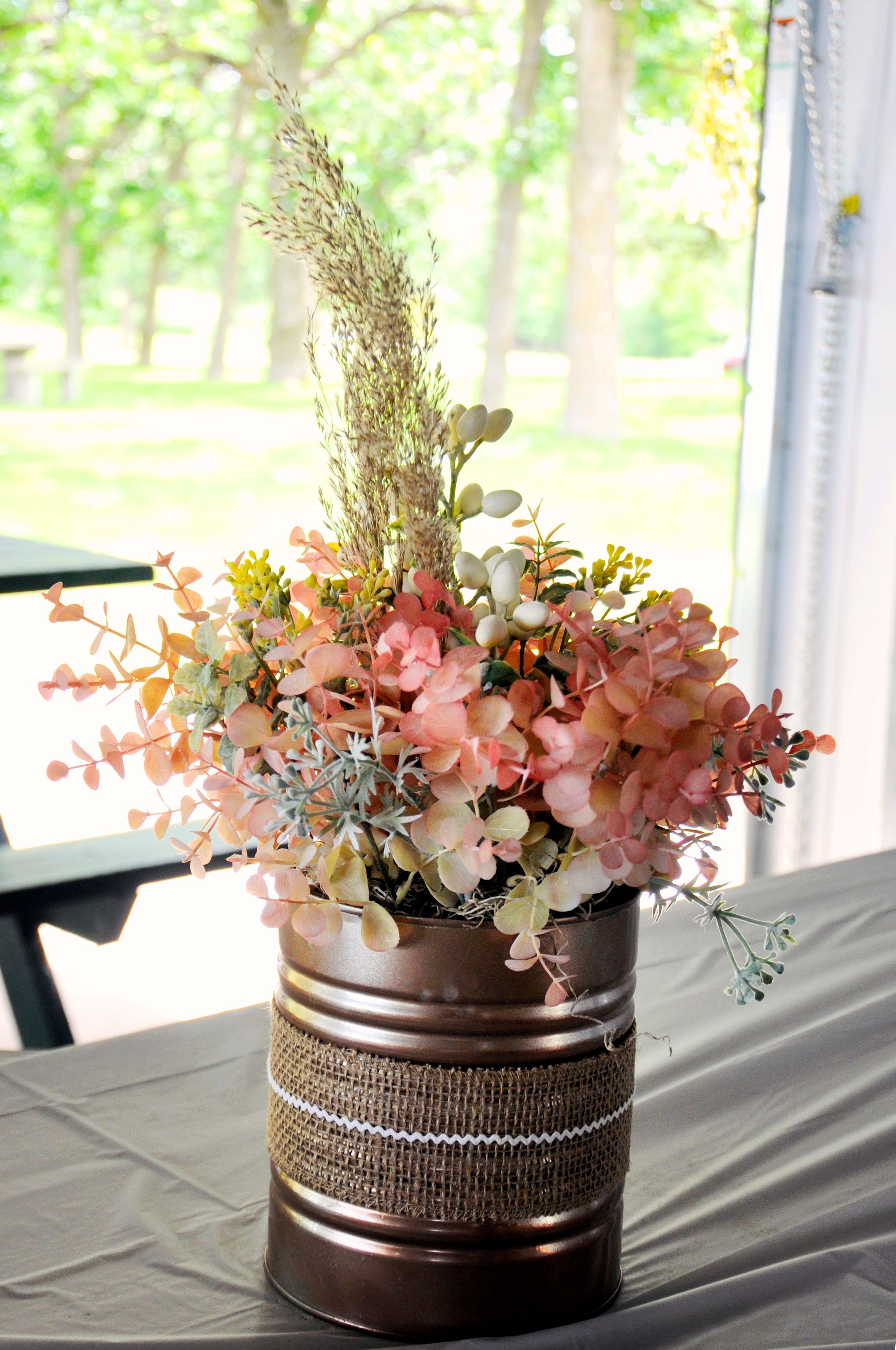 Rustic centerpiece with pink flowers and burlap ribbon wrapped around a brown container.