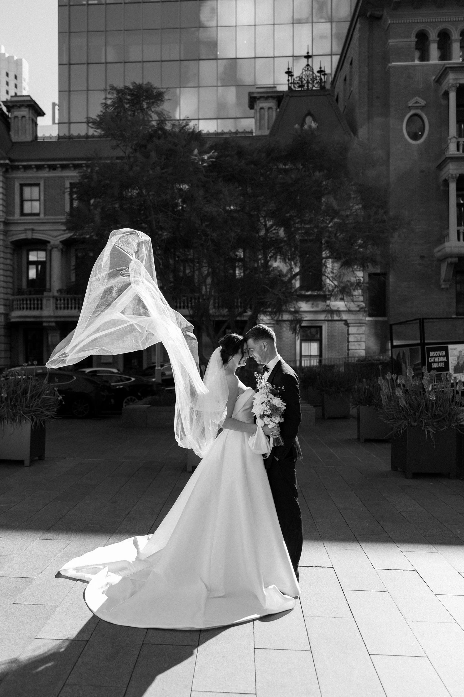 A dramatic black and white wedding photo with a flowing veil caught in the wind against an urban backdrop.