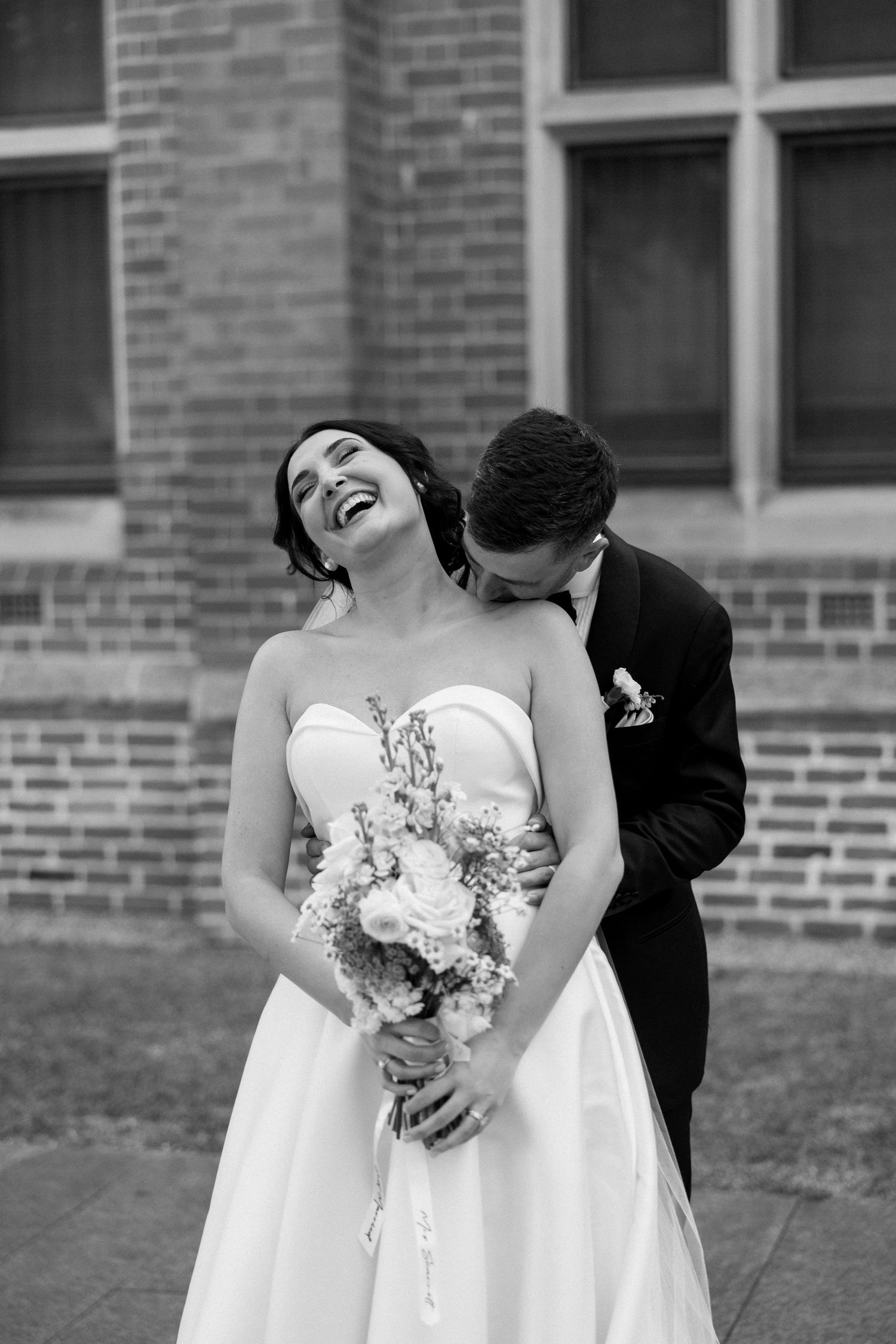A joyful candid wedding moment captured in black and white as a couple embraces while holding a floral bouquet.