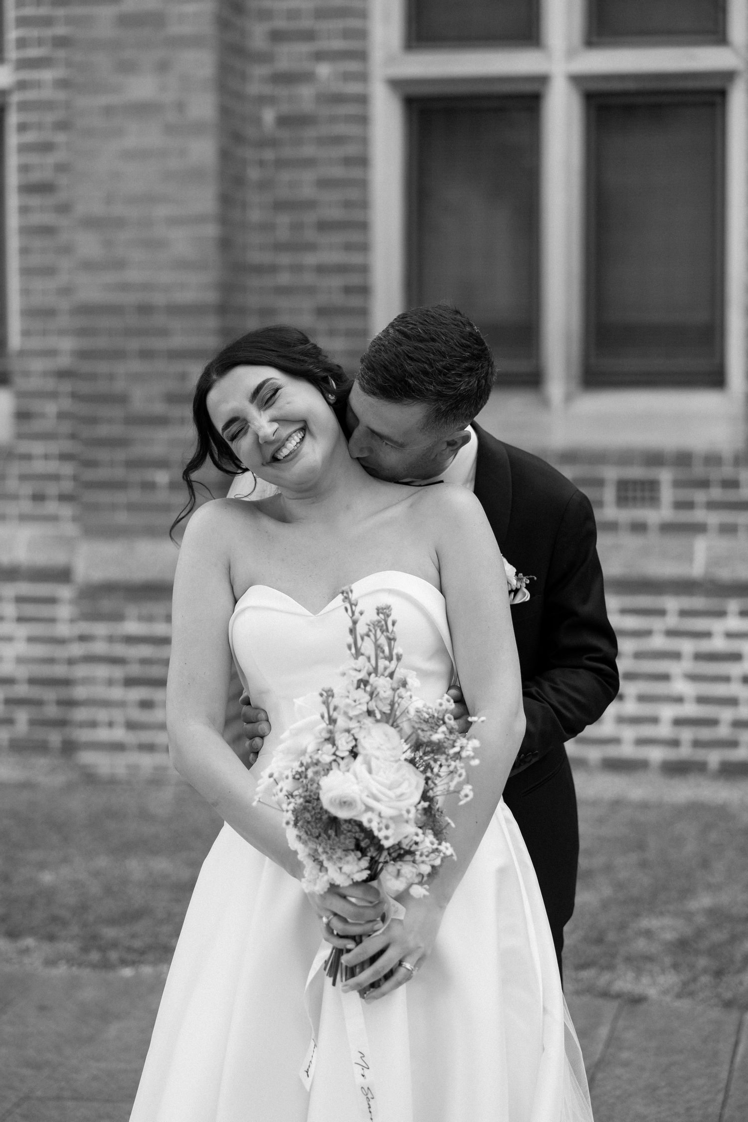 A joyful bride in a white strapless gown holds a floral bouquet while sharing an intimate moment against a brick building.