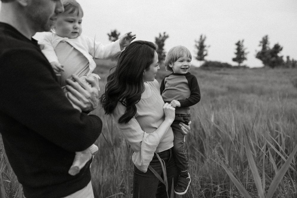 Black and white photo showing a family holding their children while standing in a field of tall grass during sunset.