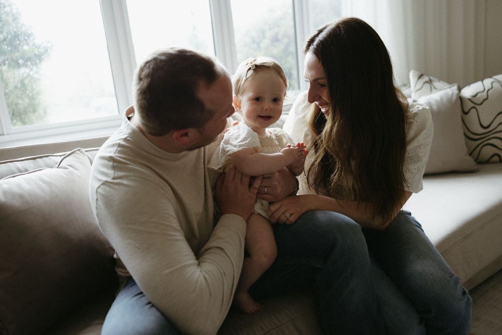 A young couple sits on a couch by a window with their baby in a tender family moment.