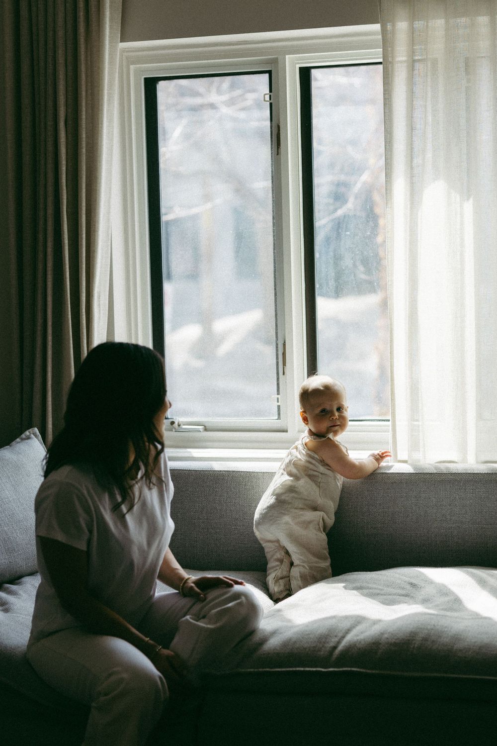 Individuals relax together on a gray couch by a sunlit window with sheer curtains in a cozy living room.