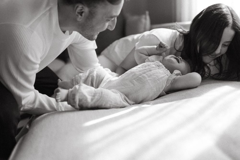 A tender black and white photo of parents lovingly gazing at their newborn baby lying on a bed.