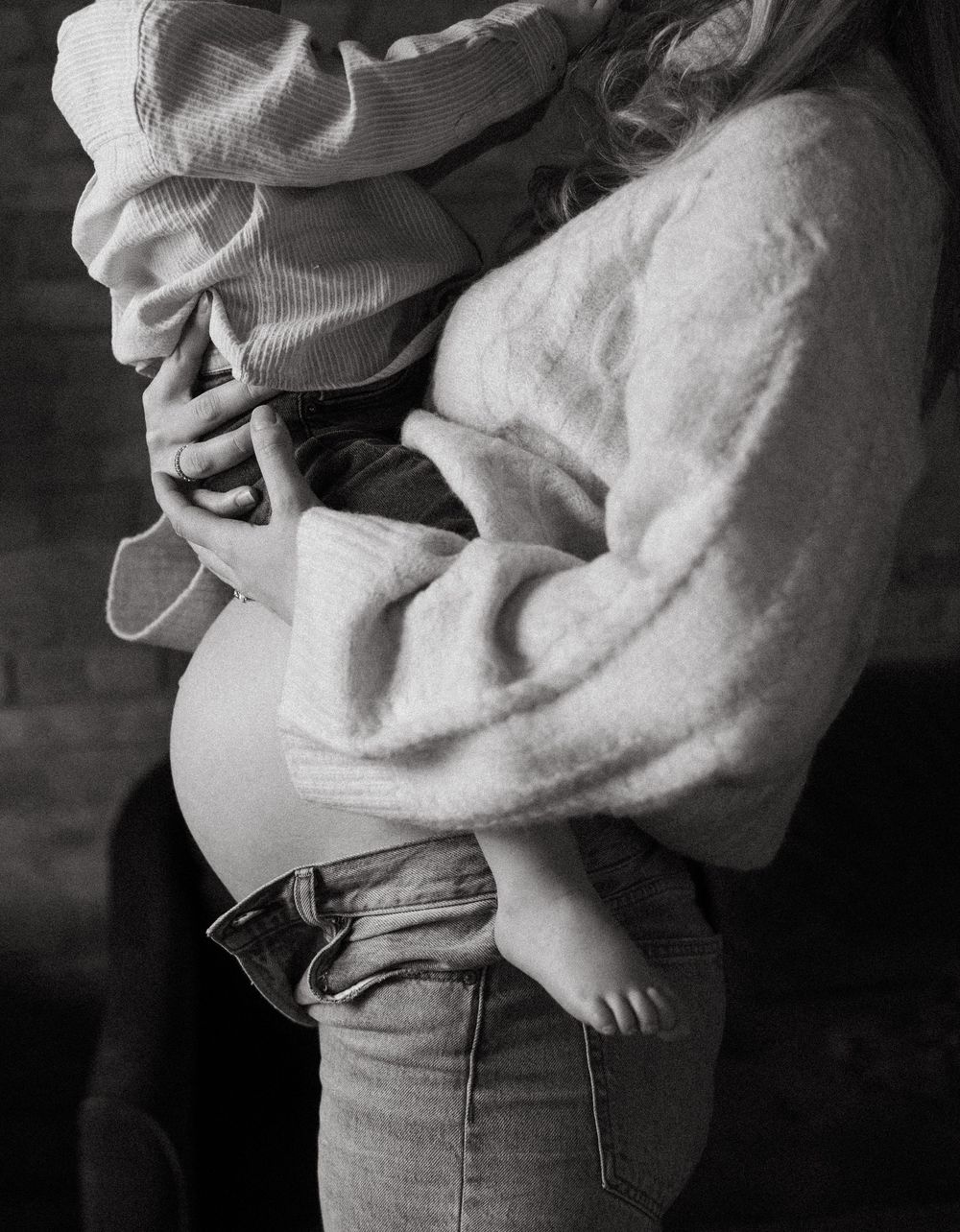 Black and white image showing a tender moment with a baby in white clothing being held against someone wearing jeans.
