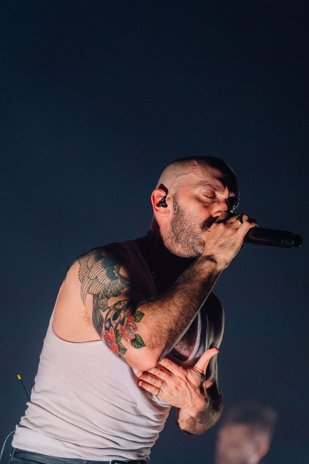 Performer in white tank top singing passionately into microphone against dark background.