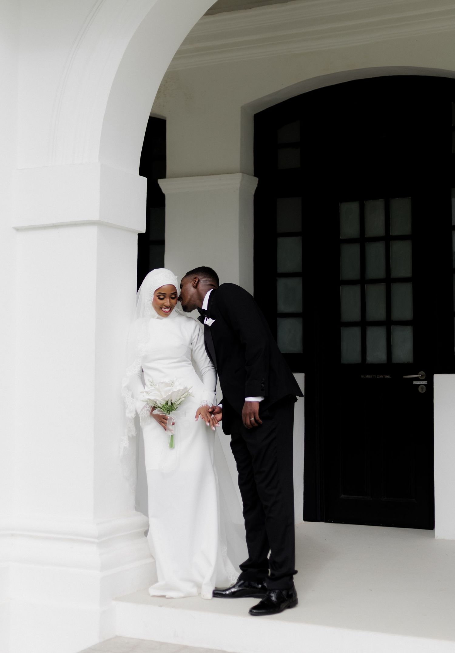 A groom in a black tuxedo kisses his bride in a white hijab and gown, holding a bouquet, at an elegant venue.
