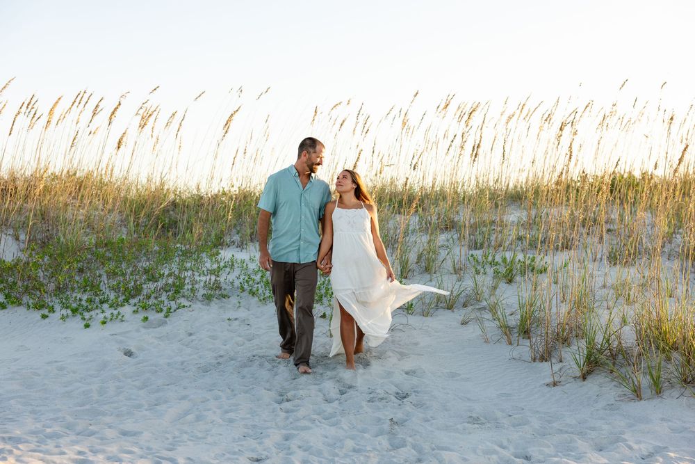 Couple walking hand in hand on white sand beach dunes with sea oats at golden hour.