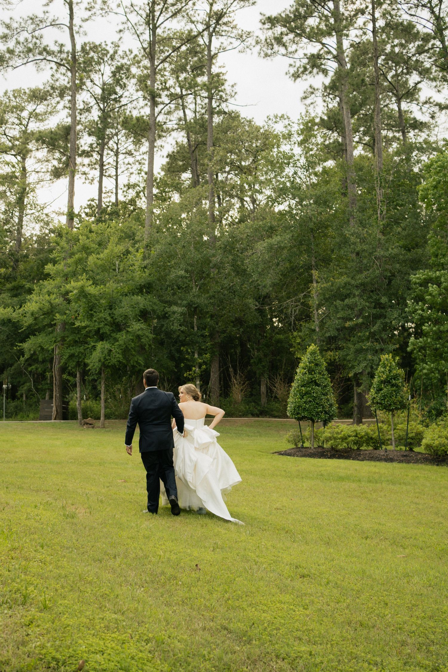 A couple in wedding attire walks hand in hand across a lush green lawn surrounded by tall pine trees.