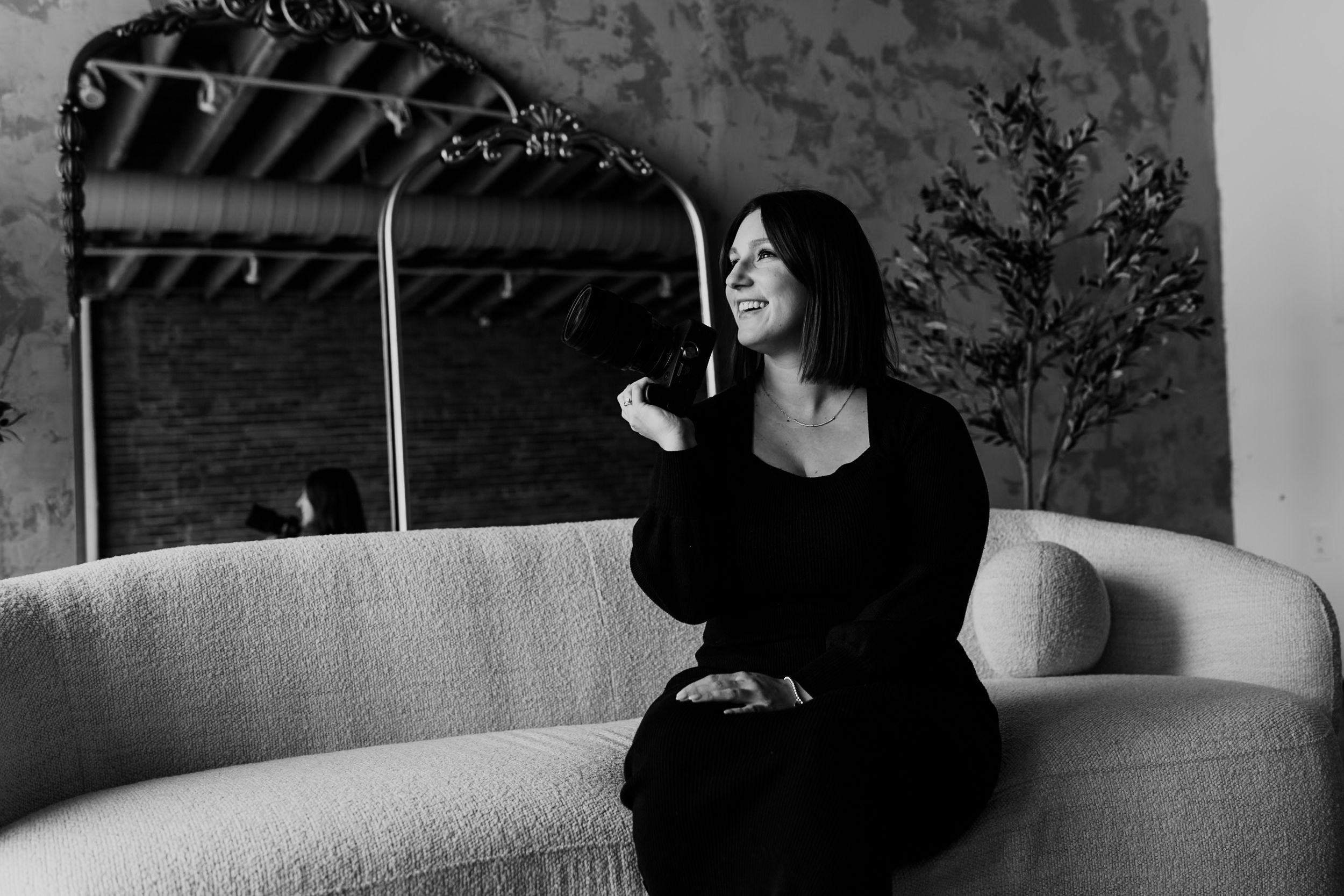 A woman in black lounges on a sofa while laughing in dramatic black and white lighting.