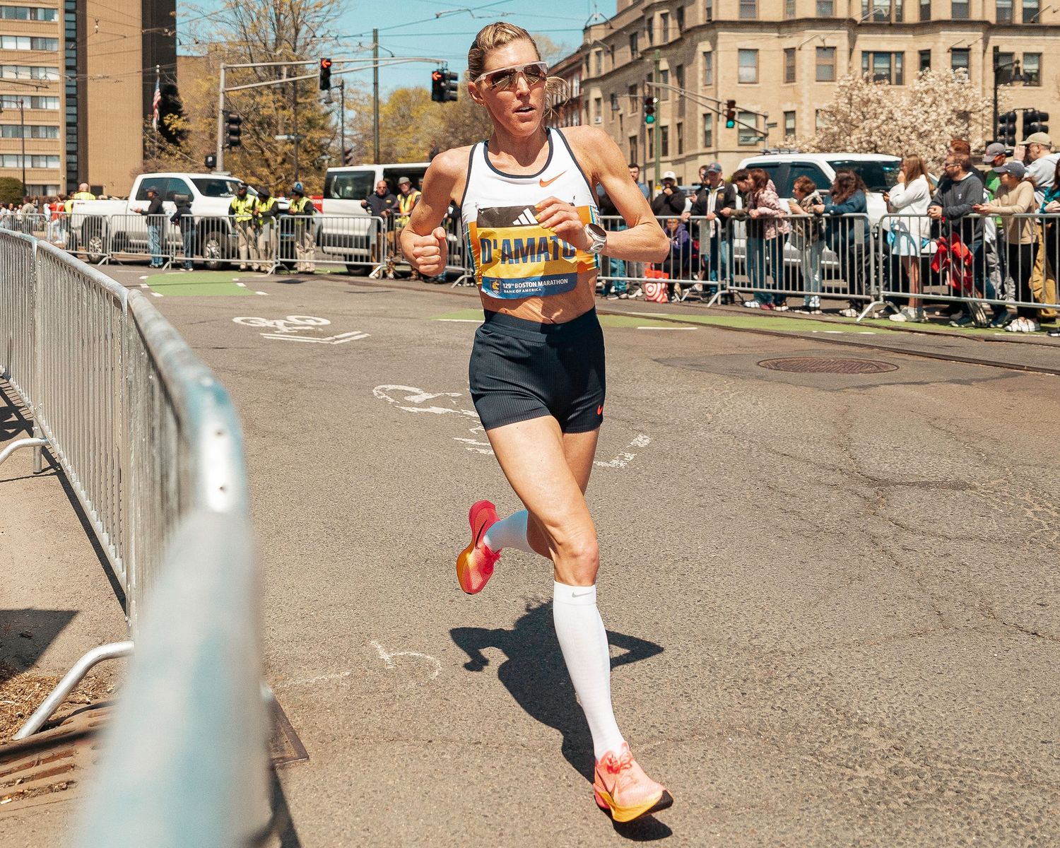 Marathon runner in competition gear races along urban course with spectators behind barriers on sunny day.