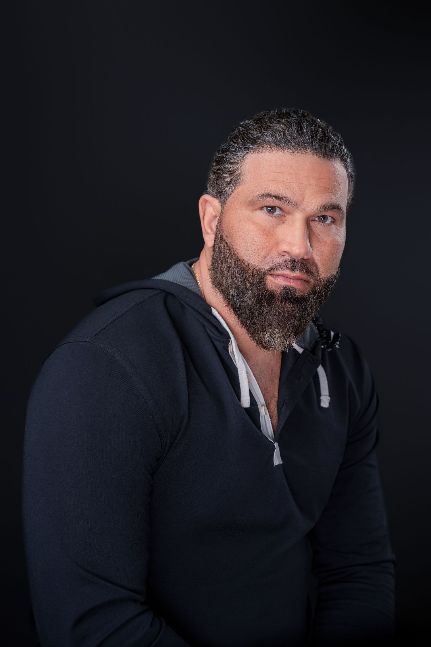 Portrait of bearded person in black hoodie against dark background.