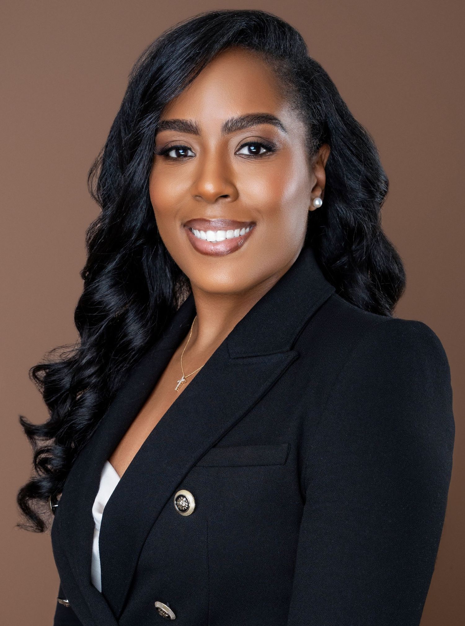 Professional headshot of person in black blazer with curled hair and bright smile against brown background.