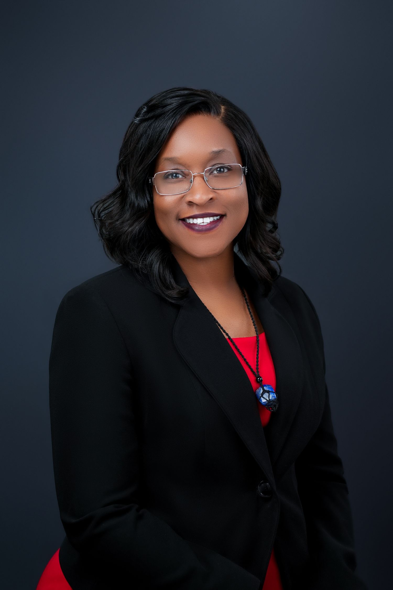 Professional portrait in black blazer with red accent against dark background.