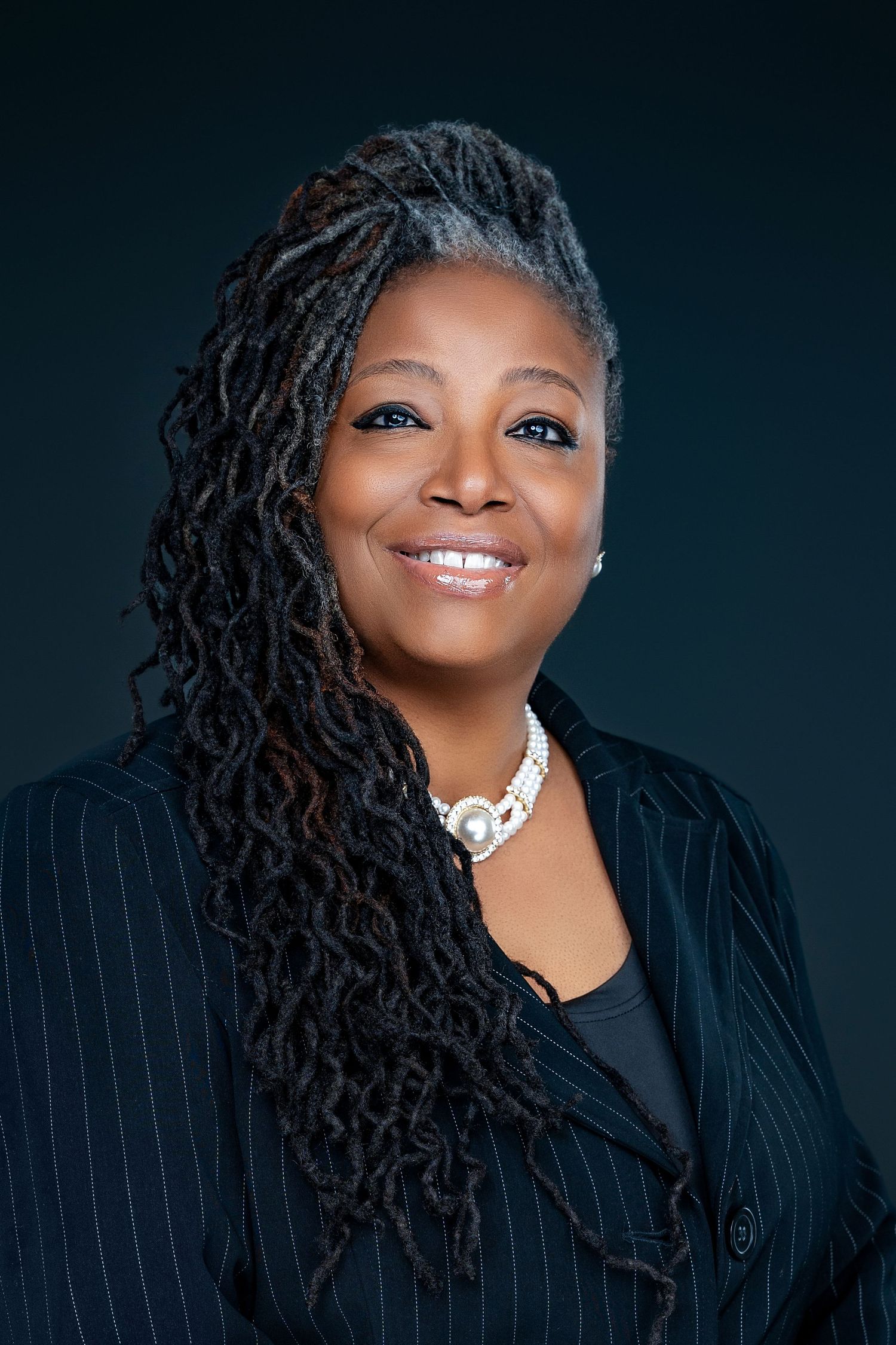 Professional portrait featuring long locs hairstyle and pinstriped black blazer with statement necklace.