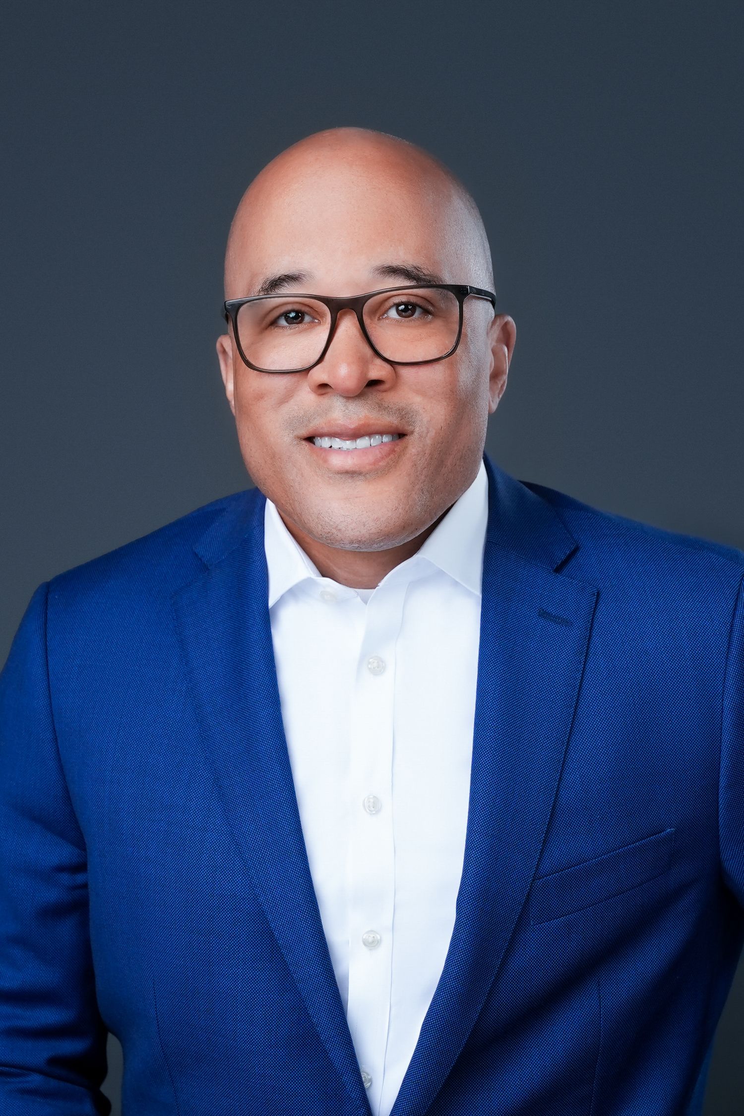 Corporate headshot of person in royal blue suit with glasses against dark background.
