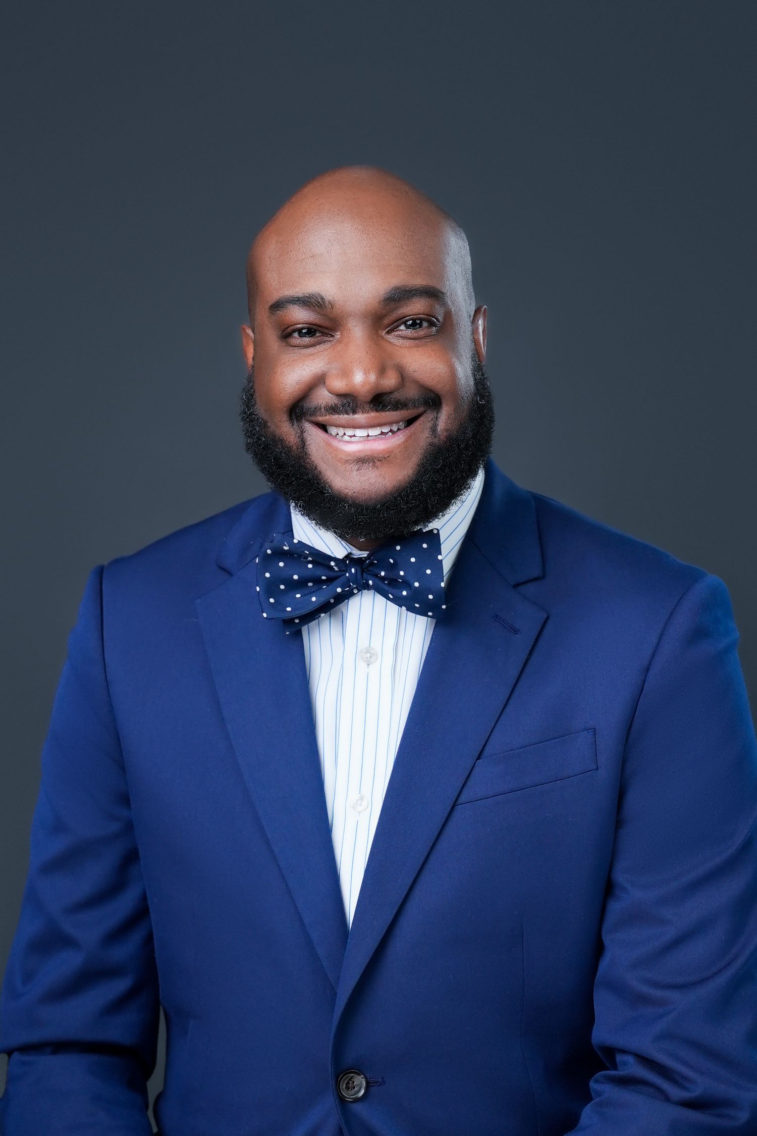 Professional portrait of person in blue suit with polka dot bowtie against dark backdrop.