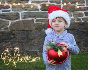 A festive scene with a red Santa hat, ornament and decorative gold 'Believe' text against a stone wall background.