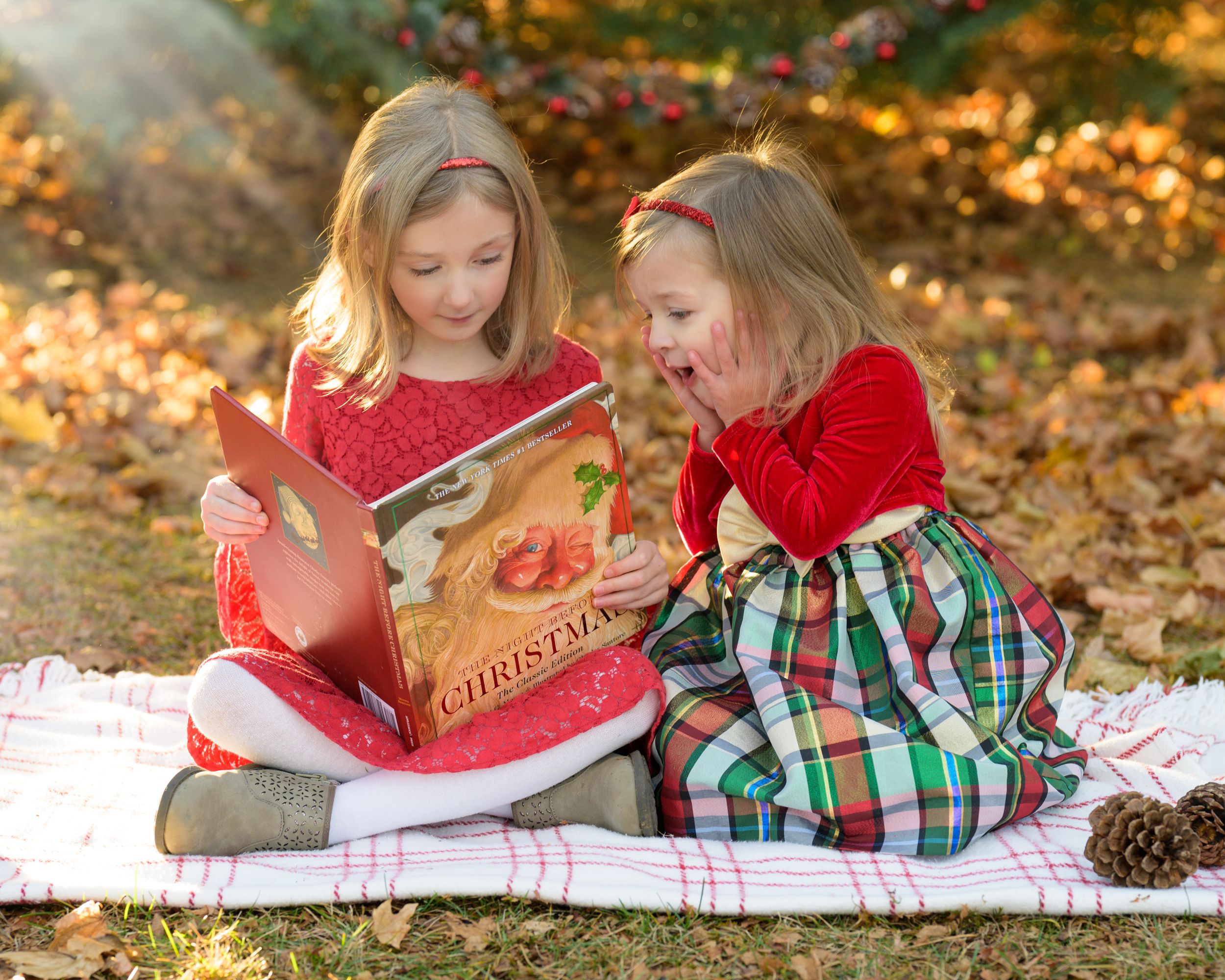Two children in holiday dresses read, The Night Before Christmas, together while sitting on a blanket surrounded by autumn leaves.