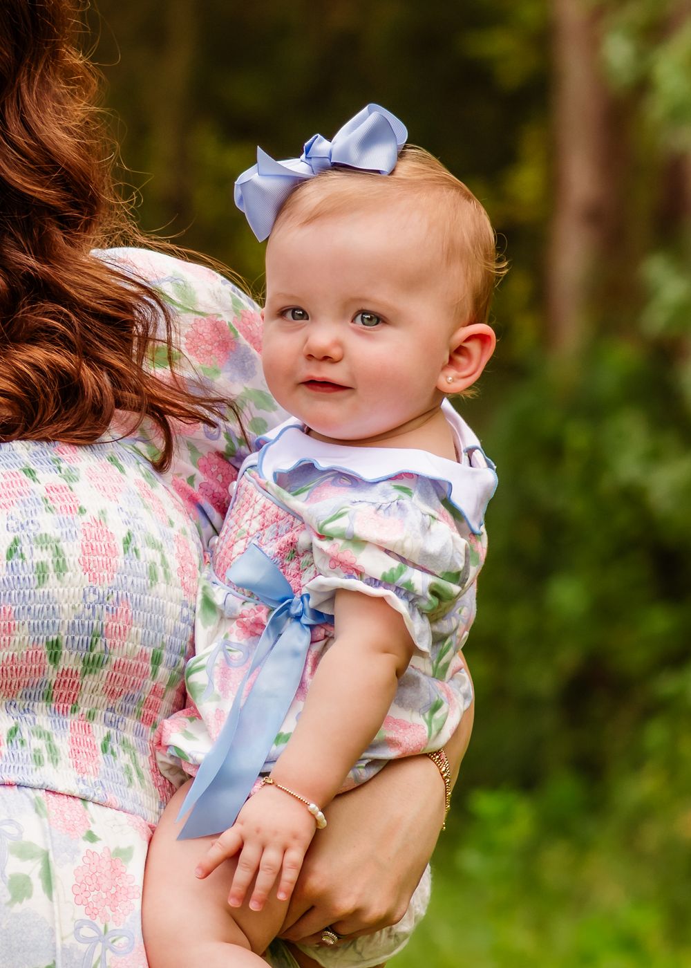 Adorable baby girl in floral dress with blue bow headband being held by mother outdoors.