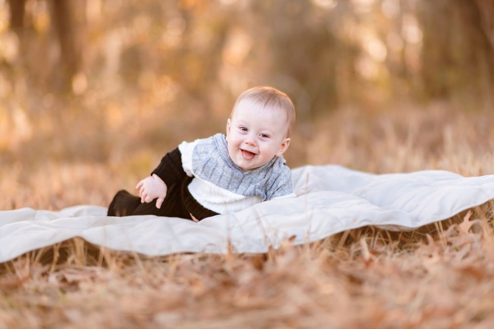 Smiling baby in a blue sweater lying on white blanket outdoors with autumn foliage in background.