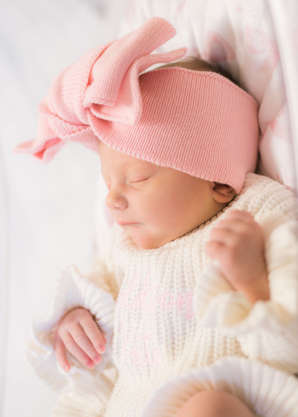 Sleeping newborn wearing white knit sweater and pink bow headband rests peacefully on white bedding.