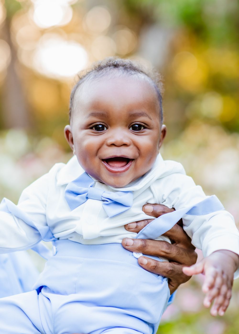 Smiling baby boy in light blue outfit held outdoors with bokeh background.