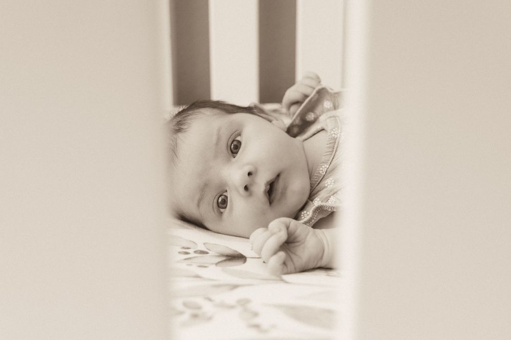 Baby lying between white crib slats in black and white.