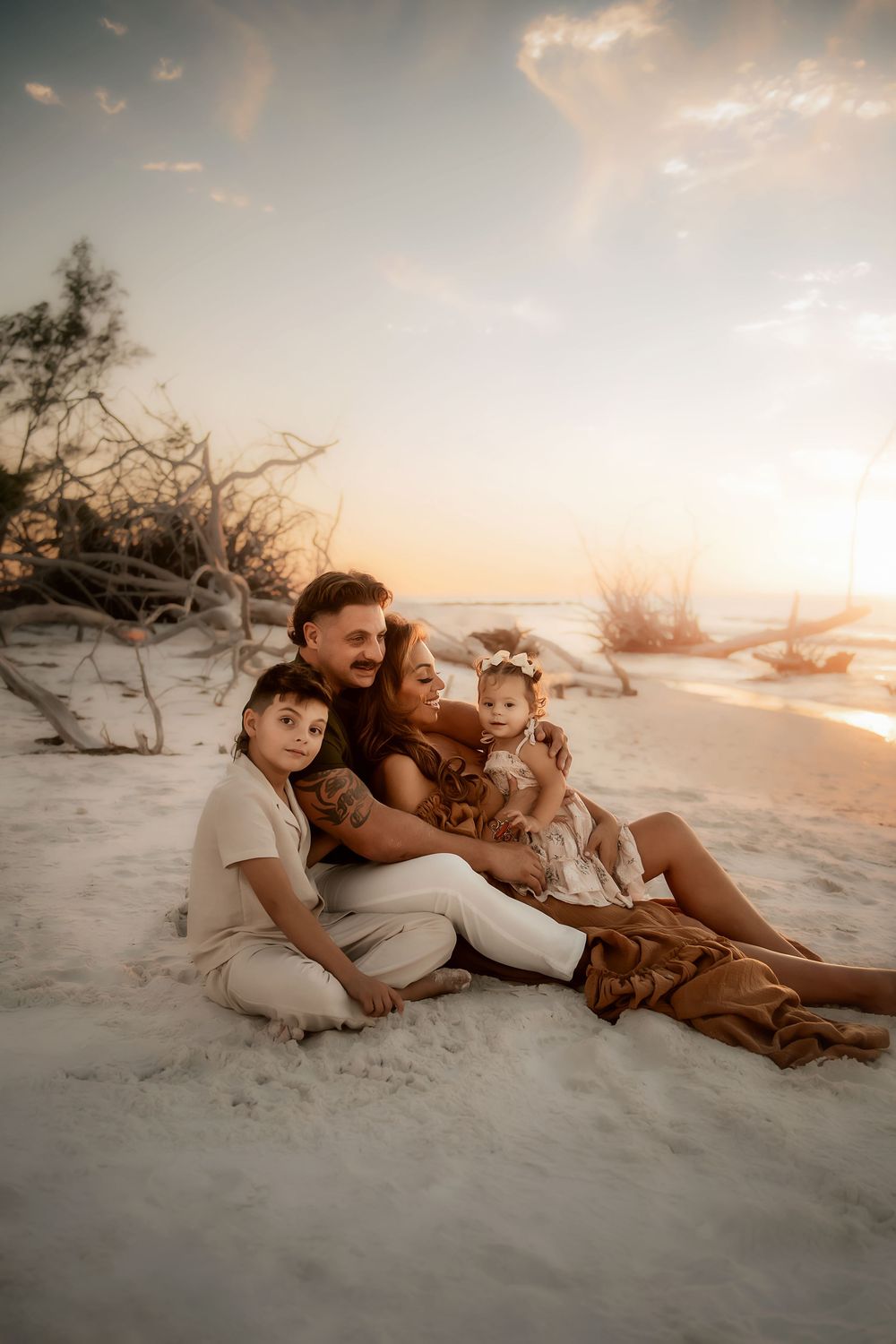 A family sits together on a sandy beach at sunset with driftwood and warm lighting creating a dreamy atmosphere.