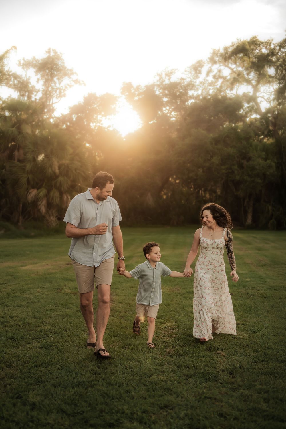 A family walks together on a grassy field during golden hour sunset with trees silhouetted in the background.