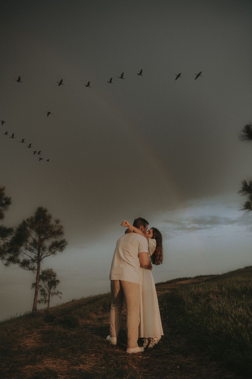 A couple stands close together as birds fly overhead in a moody sunset sky.