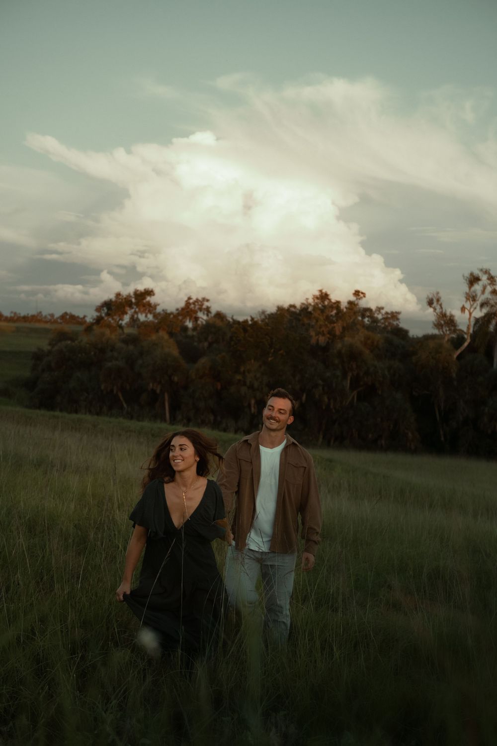Two people walk together through a serene meadow at dusk with trees silhouetted against a moody sky.