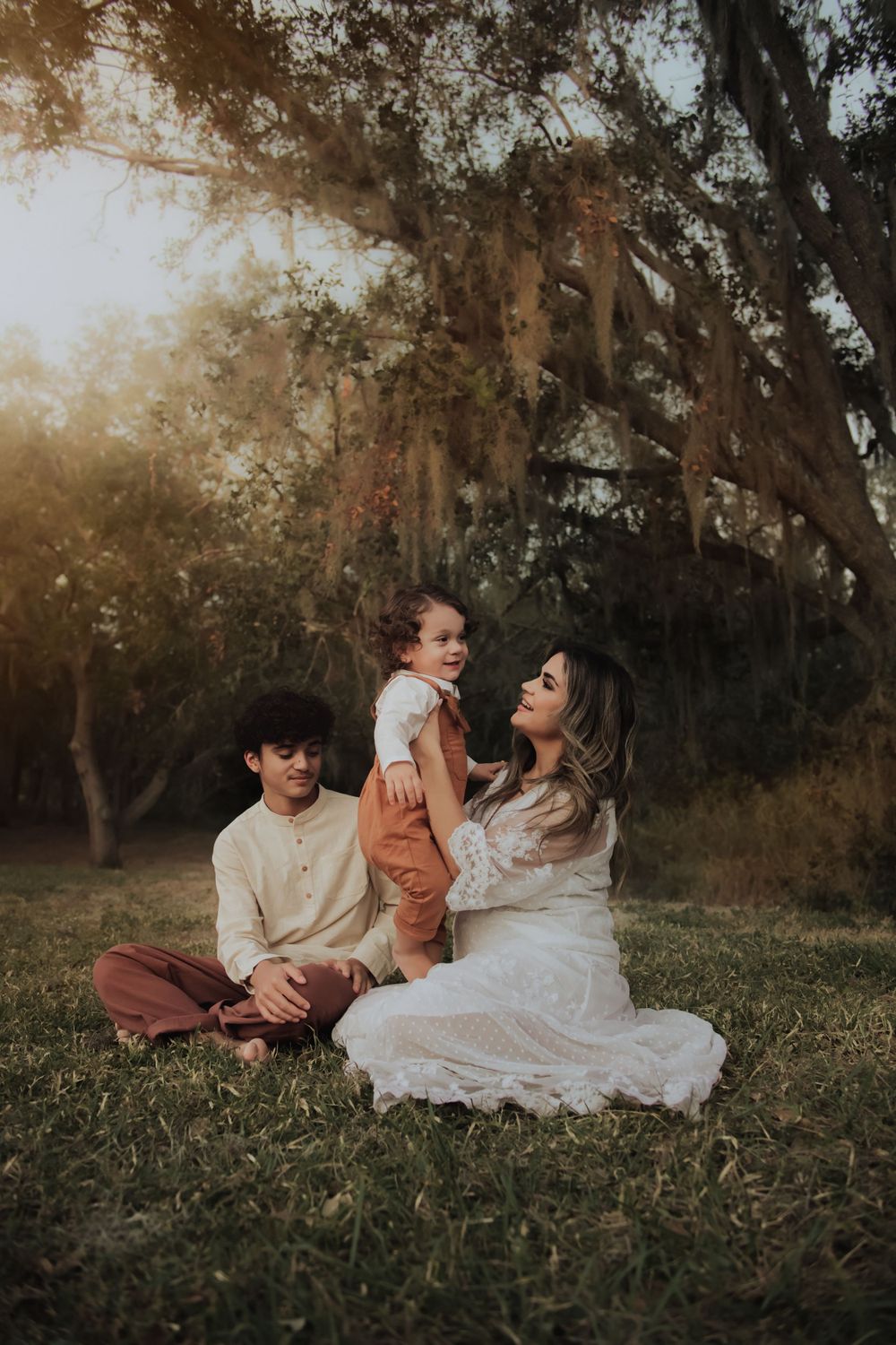 Family sitting together on grass under mossy oak trees during golden hour sunset.