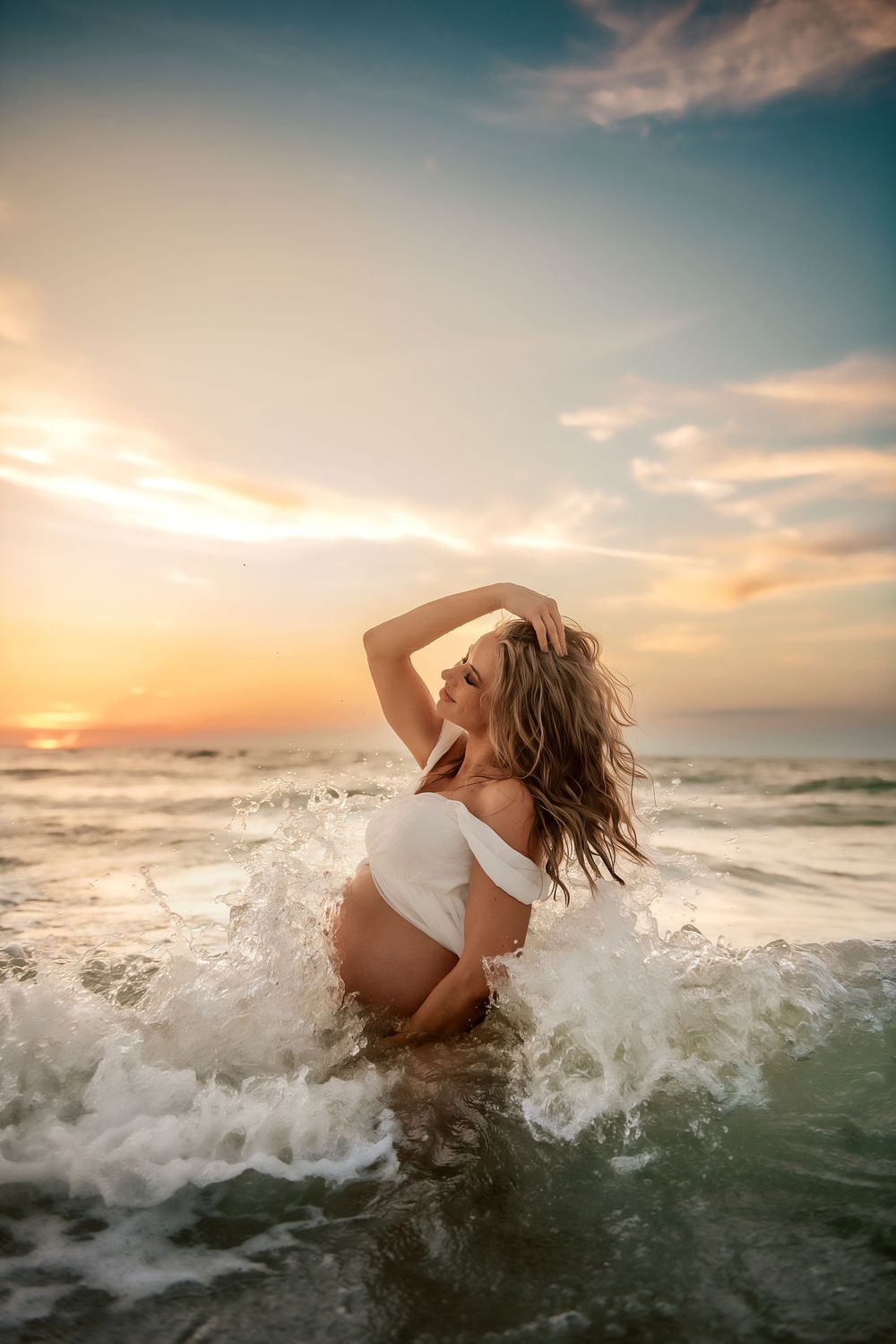 A graceful figure in white stands in ocean waves during a dramatic sunset with splashing water and golden light.