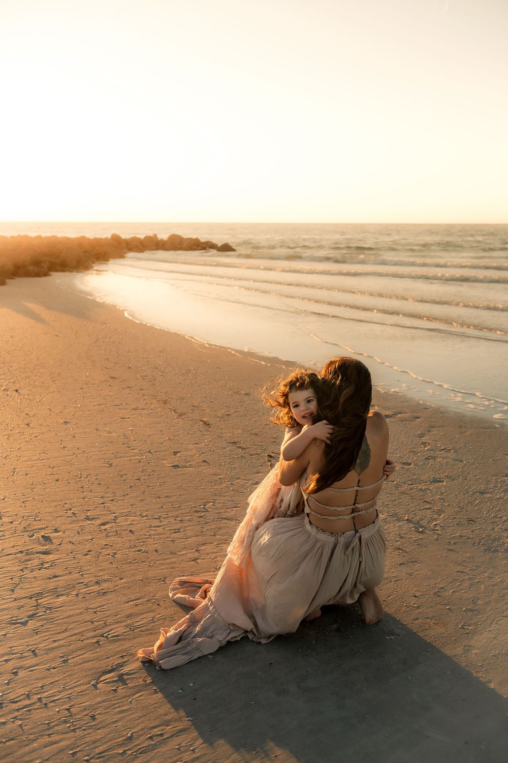 Two people share intimate moment sitting on beach sand during golden hour sunset.