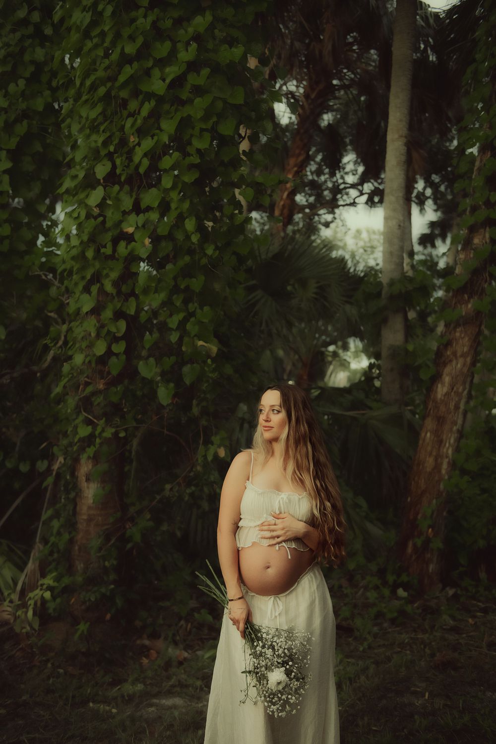 An ethereal maternity portrait set among lush green foliage and tall trees in a natural woodland setting.