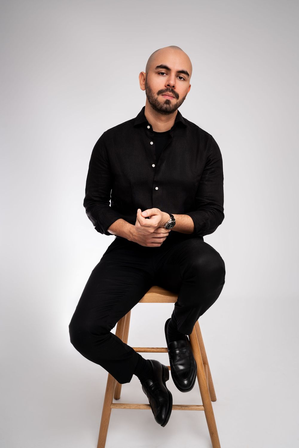 Professional studio portrait of person in all black attire sitting on wooden stool against white background. London, UK