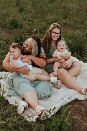 Three people sitting on blanket outdoors with babies sharing joyful moment together.