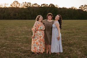 Three people wearing dresses stand together in a grassy field at sunset.