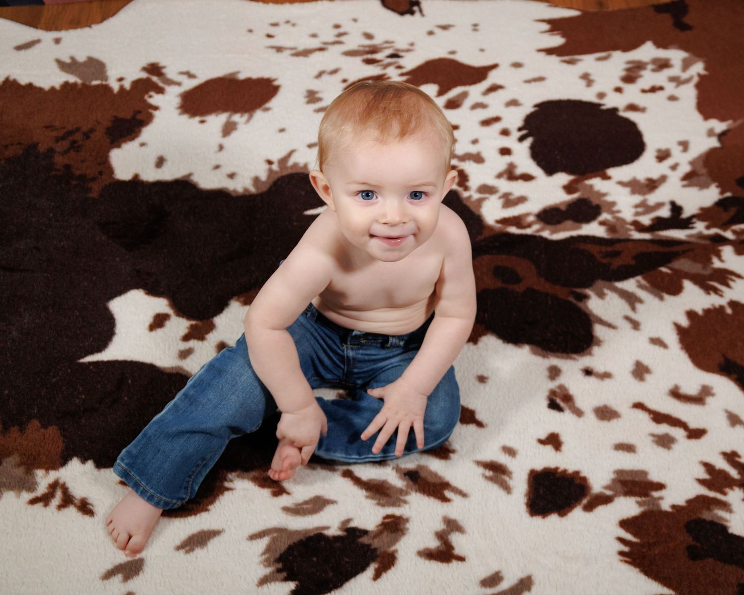 A baby wearing blue jeans sits on a cowhide rug in a studio setting.