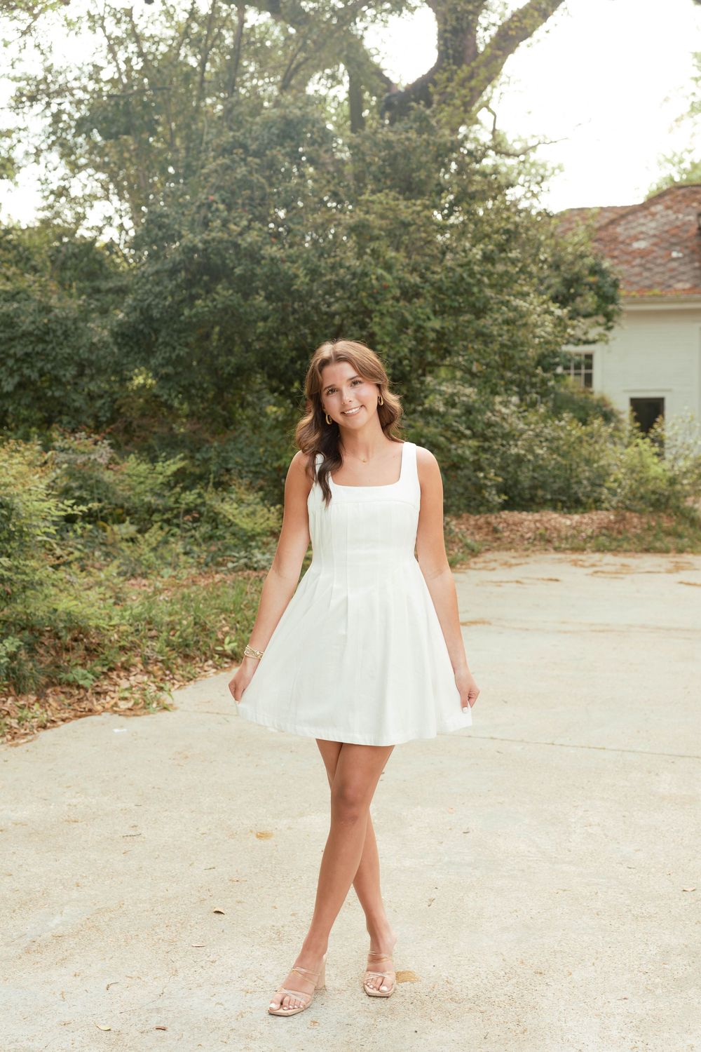 Young woman in white mini dress and nude heels posing outdoors near trees and a house.