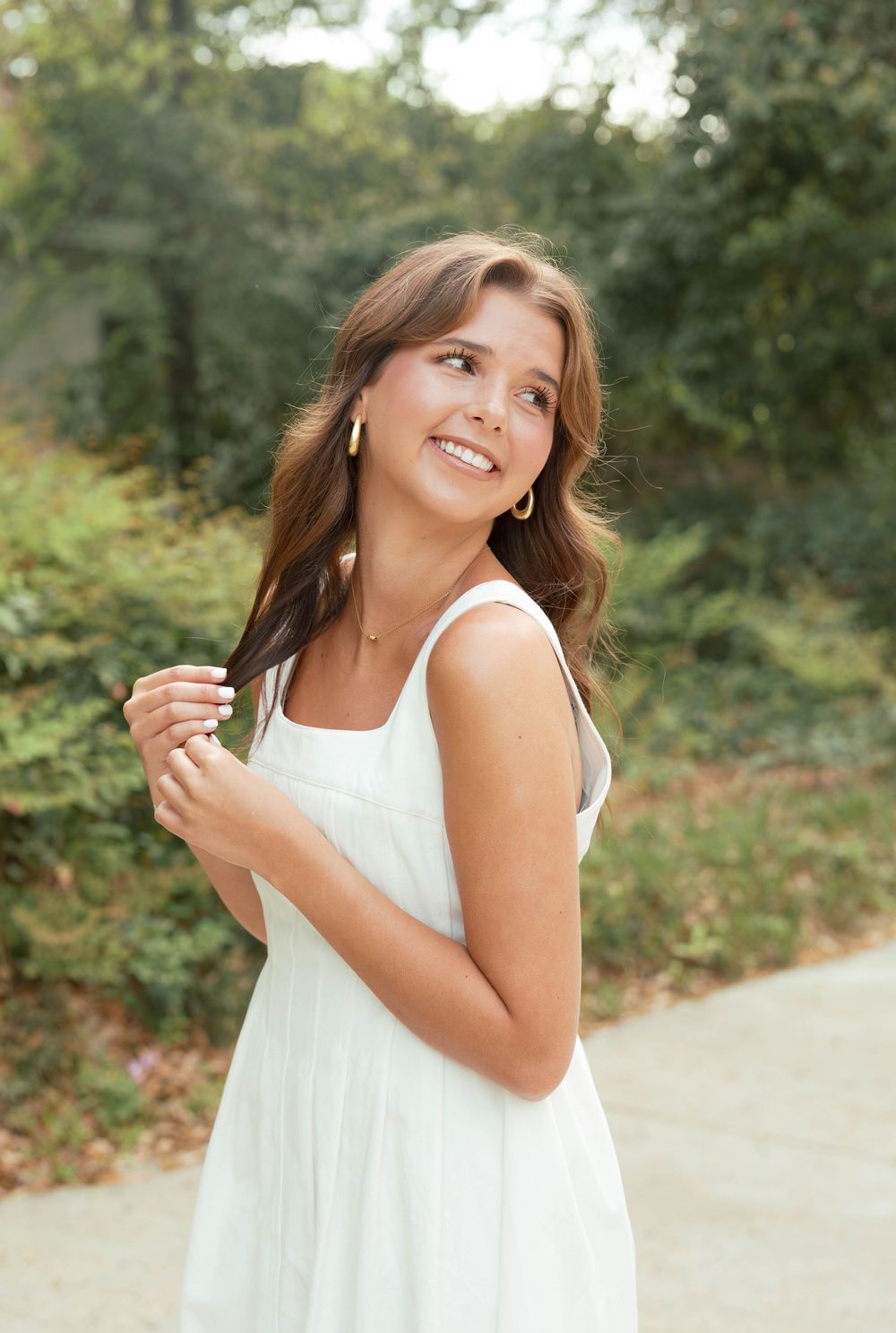 Young woman in white sundress smiling outdoors, wearing gold earrings and necklace, with lush green trees in background.