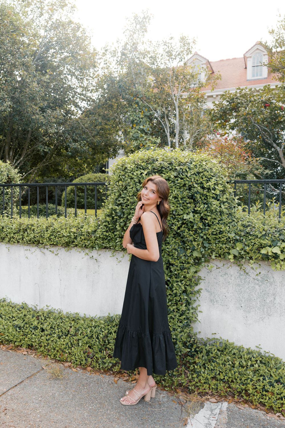 Woman in black midi dress posing outdoors by ivy-covered wall with lush green hedges and building in background.