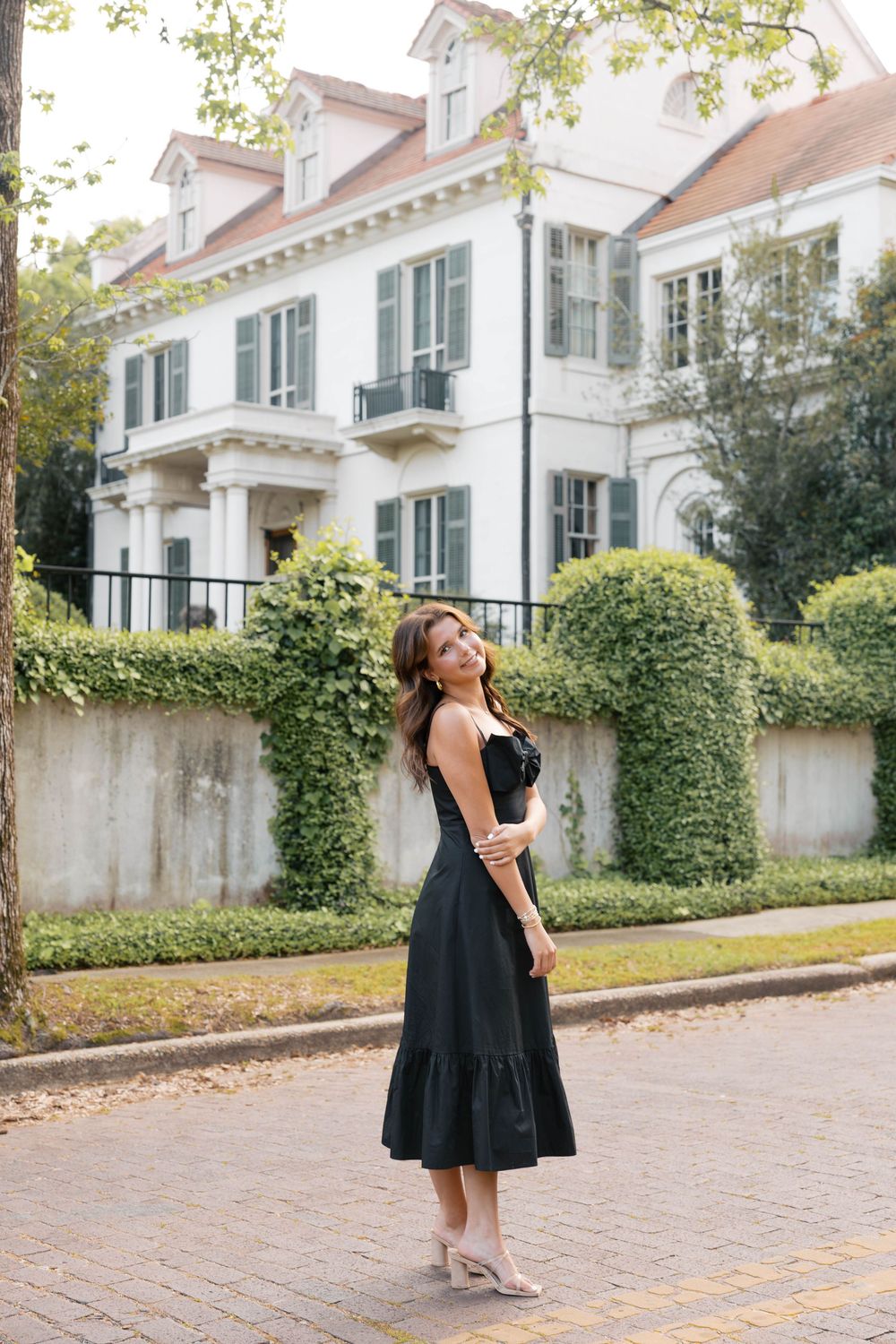 Woman in black midi dress posing on brick sidewalk in front of elegant white mansion with lush greenery.