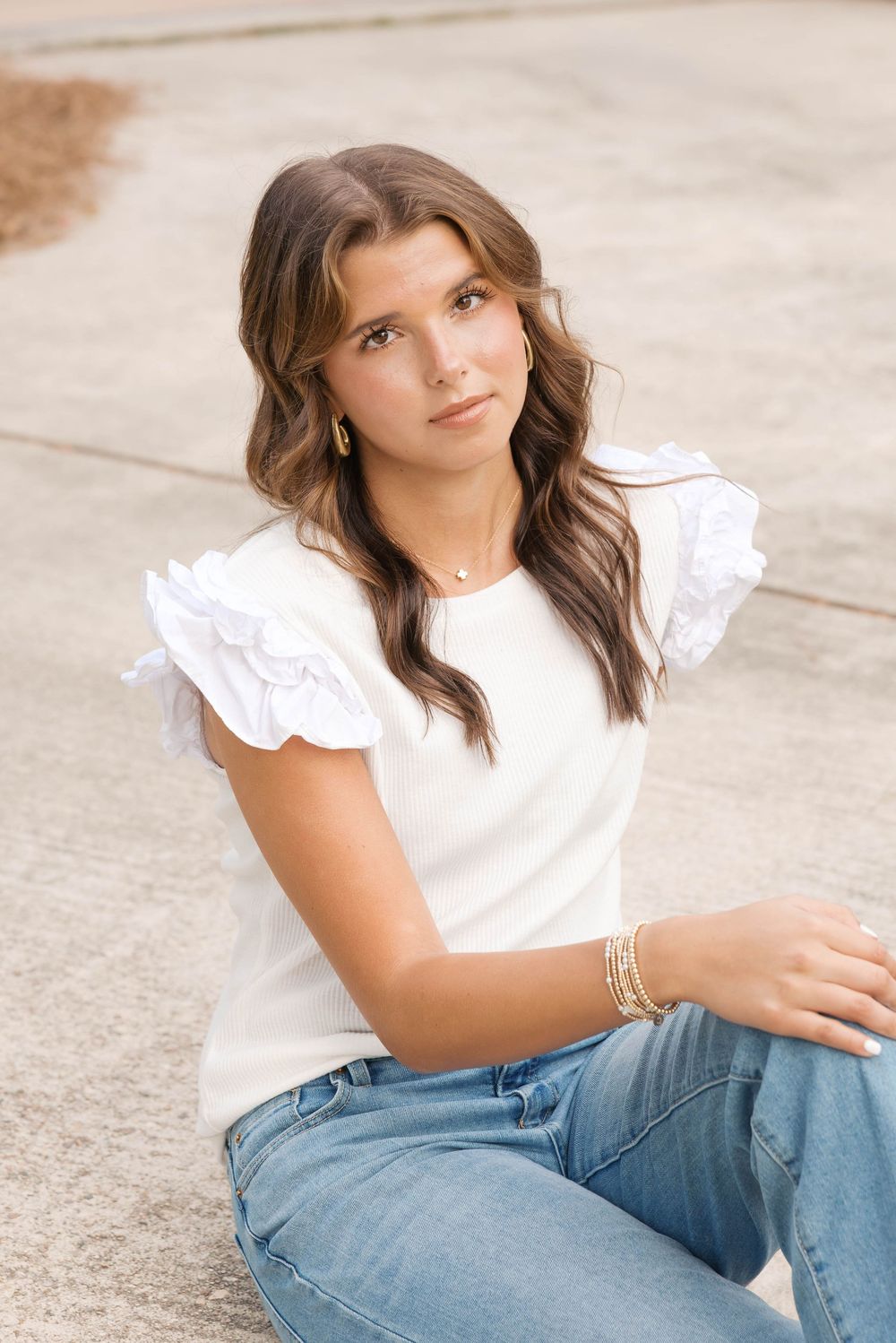 Young woman in white ruffle top and jeans sitting outdoors, wearing gold jewelry.